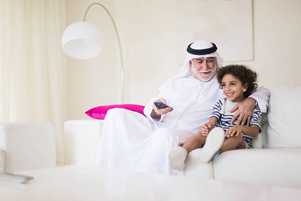 Middle eastern senior watching tv at home dressed in dishdasha.